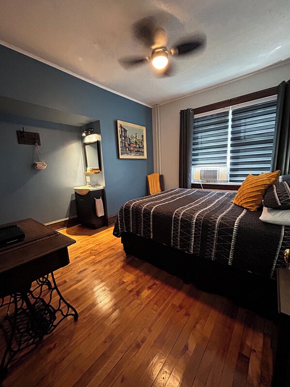 A bedroom with a bed, a desk, a chair, a ceiling fan, a window with blinds, and a painting on the wall.