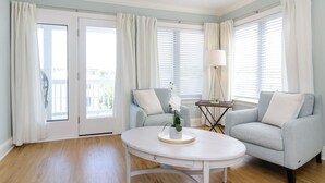 A room with a light blue wall, a white round table with a potted orchid, two light blue sofas with white cushions, a floor lamp, and a window with white curtains.