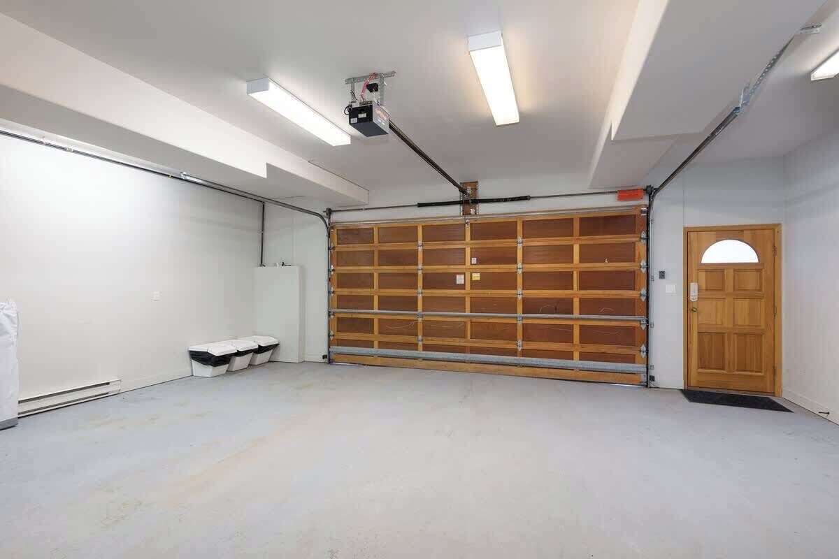 A garage with a large wooden door, fluorescent ceiling lights, and a vent on the left wall.