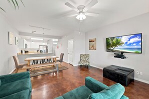 Un vaste espace de vie doté d’un ventilateur de plafond, d’une table à manger avec des chaises, d’un canapé, d’un téléviseur fixé au mur et d’une cuisine à l’arrière-plan.