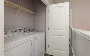 A laundry room with a washer and dryer, a wooden shelf above the washer, and a white door.