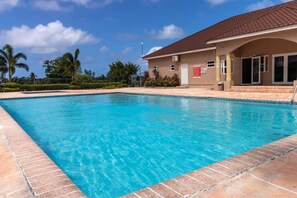 A swimming pool with a tiled deck, a building with a brown roof, and palm trees in the background.