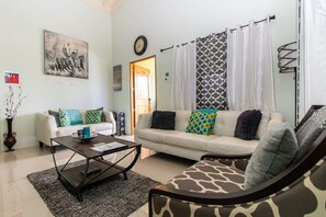 A living room with two sofas, a coffee table, a patterned armchair, a painting on the wall, and a door leading outside.