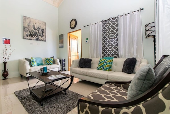 A living room with two sofas, a coffee table, a patterned armchair, a painting on the wall, and a door leading outside.