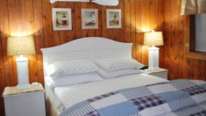 A wooden-paneled room with a bed, two bedside tables with lamps, and three framed pictures on the wall. - Beautiful Cottage Just minutes from The Twin Lakes & Beach  (Lewiston)