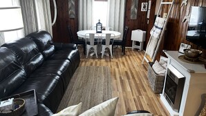 A living room with a black leather sofa, a round dining table with white chairs, a fireplace, and wooden panel walls. - Beautiful Cottage Just minutes from The Twin Lakes & Beach  (Lewiston)
