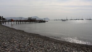 A pebbled beach with a pier extending into calm waters, boats docked in the background, and distant mountains. - Departamento, 3 dormitorios, vista al Mar (San Miguel)