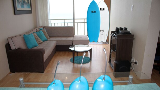 A living room with a sofa, a coffee table, and a surfboard leaning against the wall. - Departamento, 3 dormitorios, vista al Mar (San Miguel)