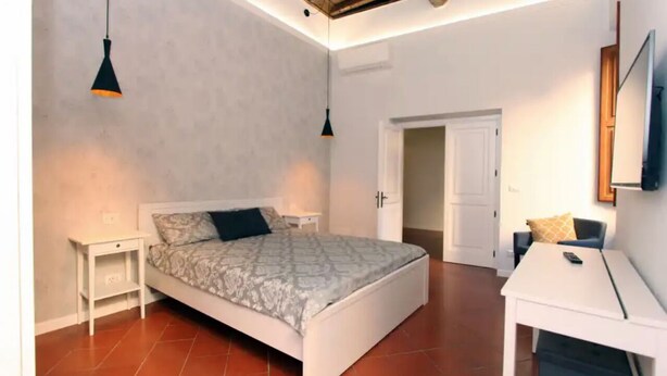 Una camera da letto con un letto, comodini, una scrivania e una sedia. Si apre una porta verso un'altra stanza.