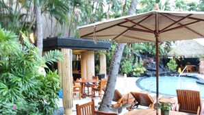A poolside area with wooden furniture, a large umbrella, and tropical plants.