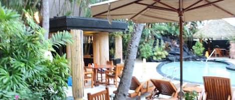 A poolside area with wooden furniture, a large umbrella, and tropical plants.