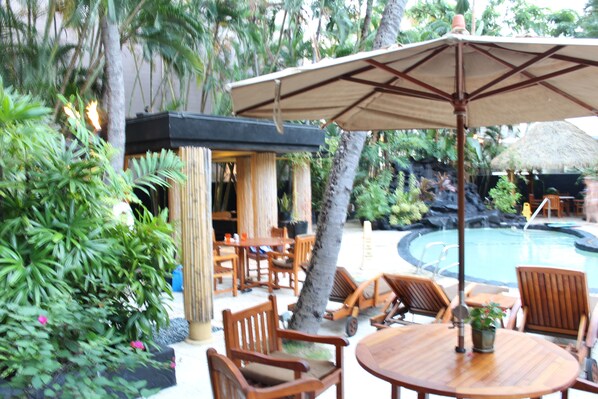 A poolside area with wooden furniture, a large umbrella, and tropical plants.