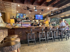 A rustic bar area with wooden furniture, ceiling fans, and a TV screen displaying a show.