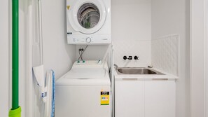 A compact laundry room with a front-loading washing machine, a small sink, and a towel rack. - Grand Pacific U104 Waterfront Views, Walk to Beach (Caloundra)