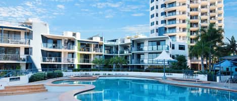 A modern hotel with a swimming pool, multiple balconies, and a clear sky.