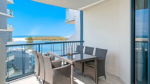 Un balcón con una mesa de vidrio y sillas de mimbre, con vistas a una playa y el océano.