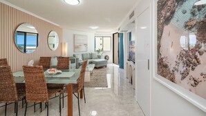 Una sala de estar moderna con un sofá, una mesa de comedor y sillas, un cuadro grande en la pared, y vistas a través de una ventana.