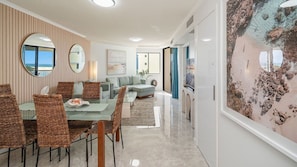 A modern living room with a sofa, dining table, and chairs, a large painting on the wall, and a view through a window.
