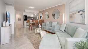 A modern living room with a sofa, coffee table, and dining area with chairs. - Grand Pacific U104 Waterfront Views, Walk to Beach (Caloundra)