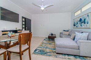 Un salon comprenant un canapé, une table basse, une table à manger avec des chaises, un ventilateur de plafond et un téléviseur fixé au mur.