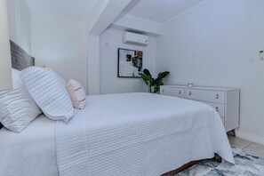 Une chambre à coucher comprenant un lit, une table de chevet, une commode, un climatiseur fixé au mur et un tableau encadré.