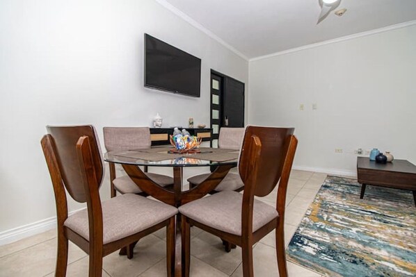Un coin repas comprenant une table ronde en verre, quatre chaises en bois, un téléviseur à écran plat fixé au mur et une petite table d’appoint agrémentée d’objets décoratifs.
