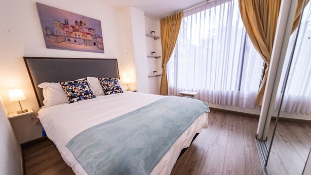 A hotel room with a large bed, a headboard, a painting on the wall, a window with curtains, and a bedside table with a lamp. - Modern Loft in Chapinero Alto (Bogotá)