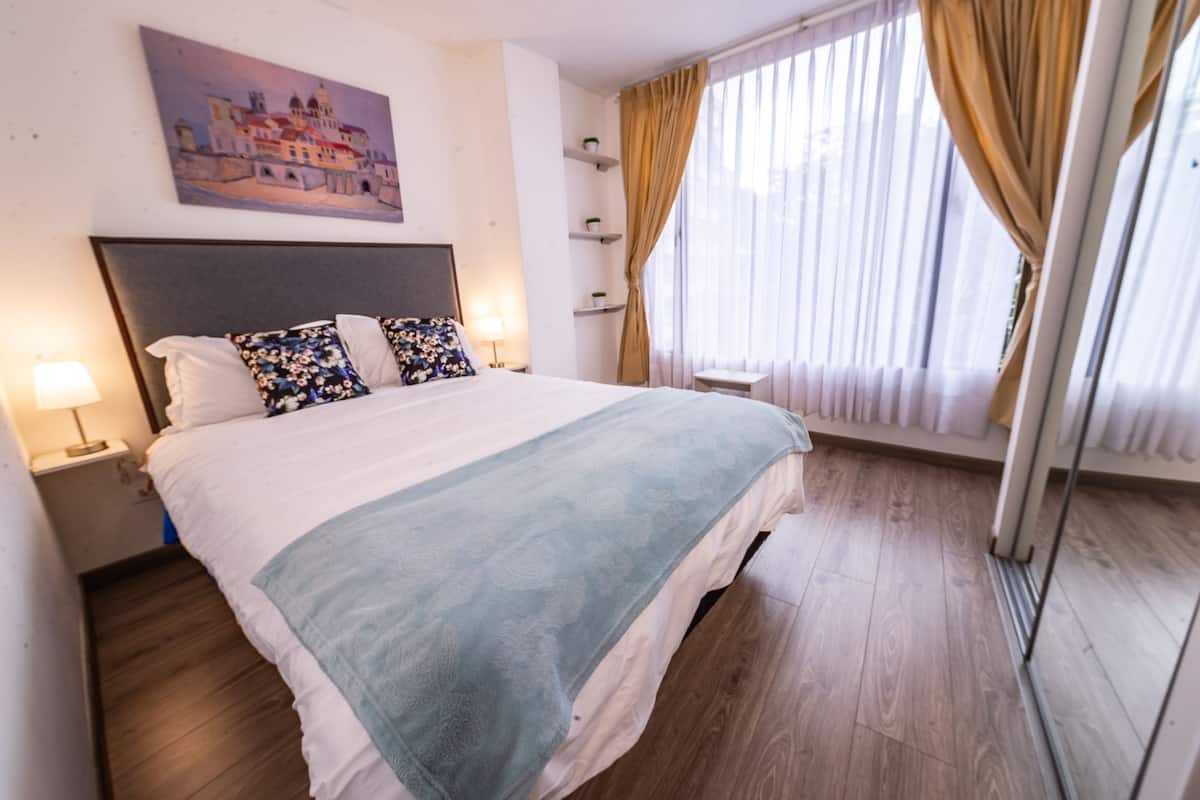 A hotel room with a large bed, a headboard, a painting on the wall, a window with curtains, and a bedside table with a lamp.