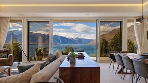 A modern living room with a large wooden table, comfortable seating, and a panoramic mountain and lake view through floor-to-ceiling windows.