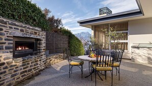 Une cheminée en pierre avec un feu, un espace extérieur aménagé avec des chaises et une table, et une vue sur les montagnes en arrière-plan.