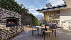 Une cheminée en pierre avec un feu, un espace extérieur aménagé avec des chaises et une table, et une vue sur les montagnes en arrière-plan.