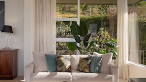 Een woonkamer met een bank, kussens, een lamp en een groot raam met planten.