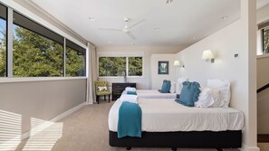 Une chambre d’hôtel avec deux lits, un ventilateur de plafond, de grandes fenêtres, une vue sur les arbres et une petite table avec une chaise.