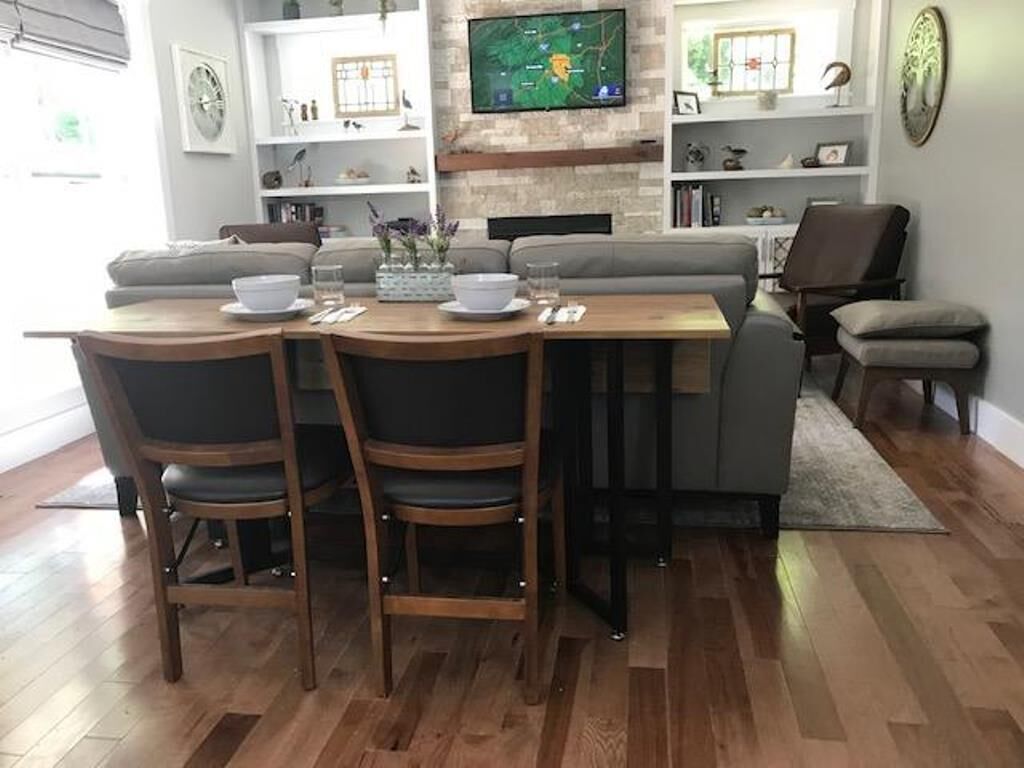 Zona pranzo con tavolo e sedie in legno, televisore a parete e divano sullo sfondo.