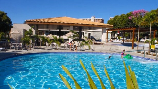 A swimming pool with clear blue water, surrounded by lounge chairs and umbrellas, with a building in the background. - International Resort (Chácara Roma)