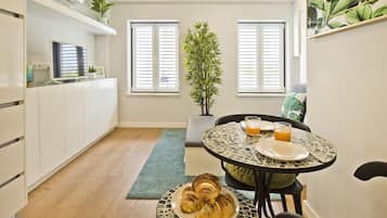 A modern kitchen with a dining area, featuring a round table with a patterned tablecloth, a bench, and a breakfast setting with croissants and orange juice.