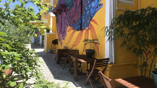 A courtyard with a mural of a fish on a yellow wall, wooden tables and chairs, and potted plants. - Beautiful Apartment at Miramar (Santurce)