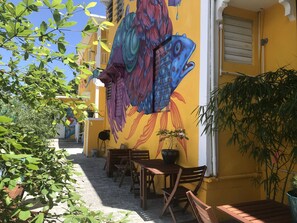 A courtyard with a mural of a fish on a yellow wall, wooden tables and chairs, and potted plants.