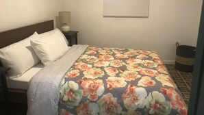A bedroom with a bed, a nightstand, a lamp, a window with blinds, and a trash can. - Beautiful Apartment at Miramar (Santurce)