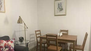 A small, neatly arranged room with a wooden dining table, four chairs, a black sofa, a small side table with a lamp, and a framed botanical illustration on the wall. - Beautiful Apartment at Miramar (Santurce)