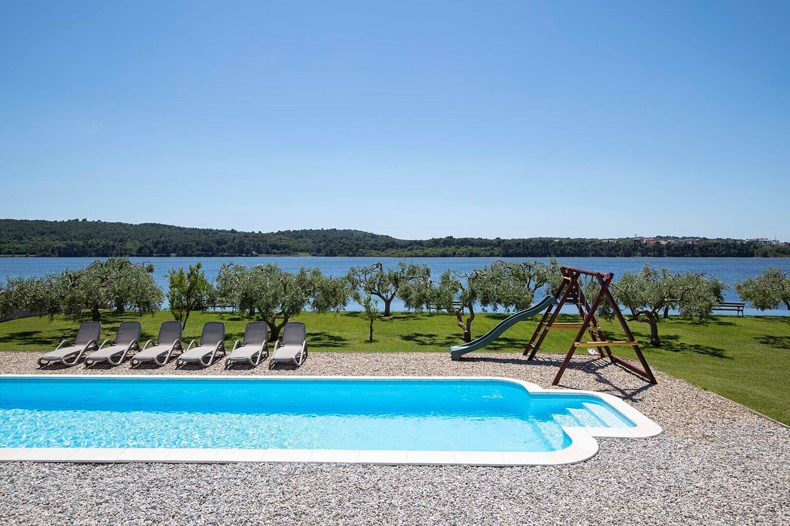 Ein Schwimmbad mit Liegestühlen und einer Rutsche, umgeben von Bäumen und Blick auf einen See.