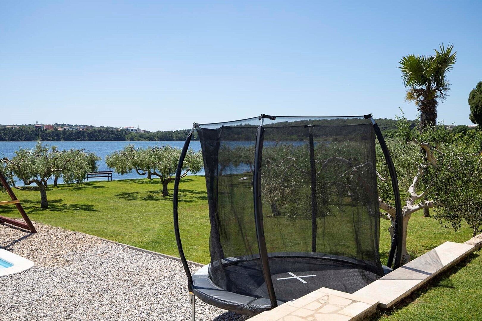 A trampoline with a safety net, surrounded by a gravel area and a grassy lawn with trees and a bench.