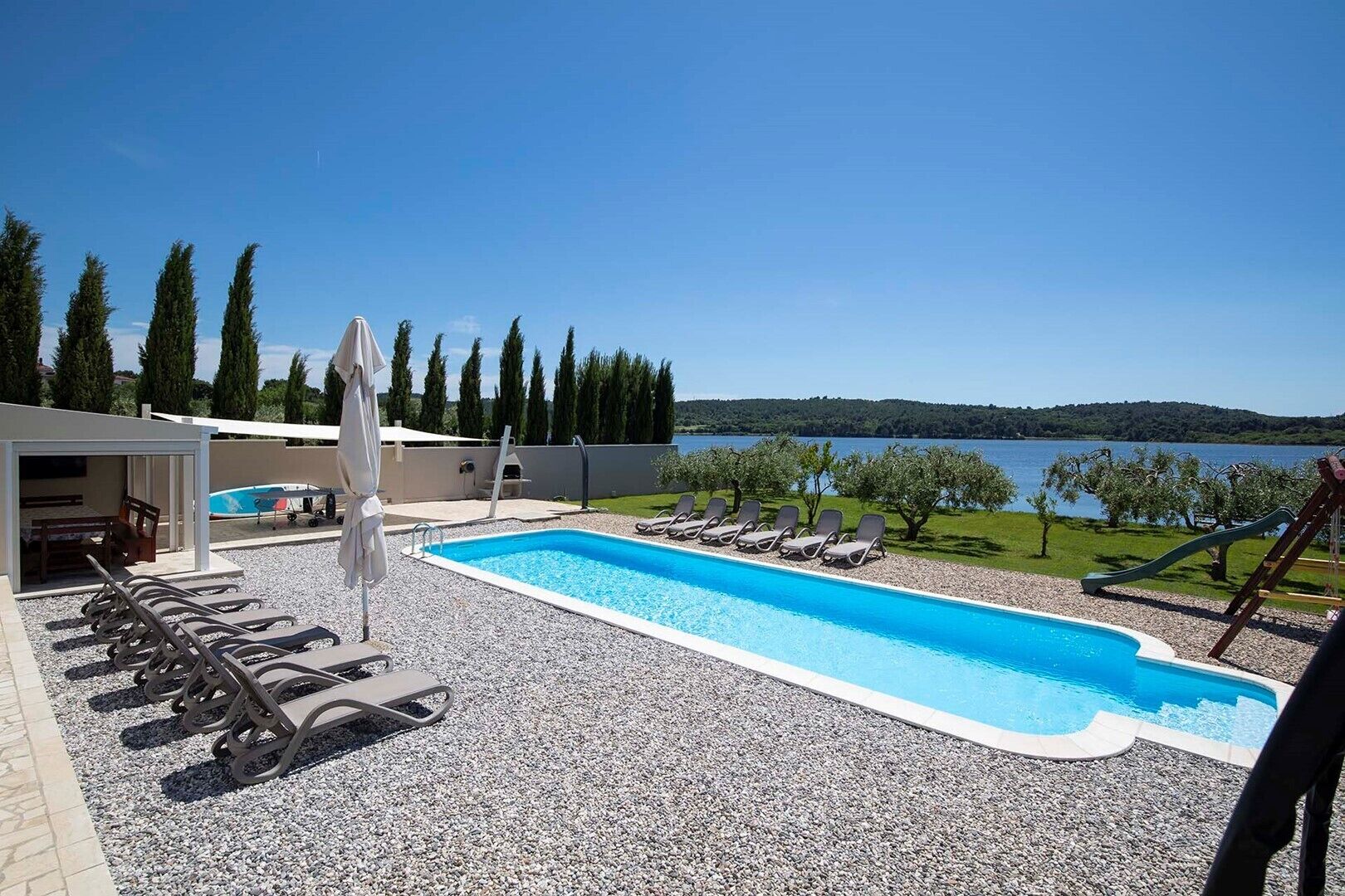 A swimming pool with lounge chairs, a covered seating area, and a view of a lake and hills.