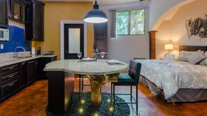 Een moderne keuken met donkerhouten kasten, een granieten aanrechtblad en een centraal eiland. Er is een zithoek met een ronde tafel en stoelen, en een slaapkamer met een bed, een nachtkastje en een wandlamp.