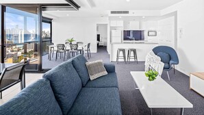 A modern apartment with a living room, dining area, and kitchen. It features a large window with a city view, a blue sofa, a dining table, and a kitchen with white cabinets and a central island. - Circle on Cavill Ocean & River Views (Surfers Paradise)
