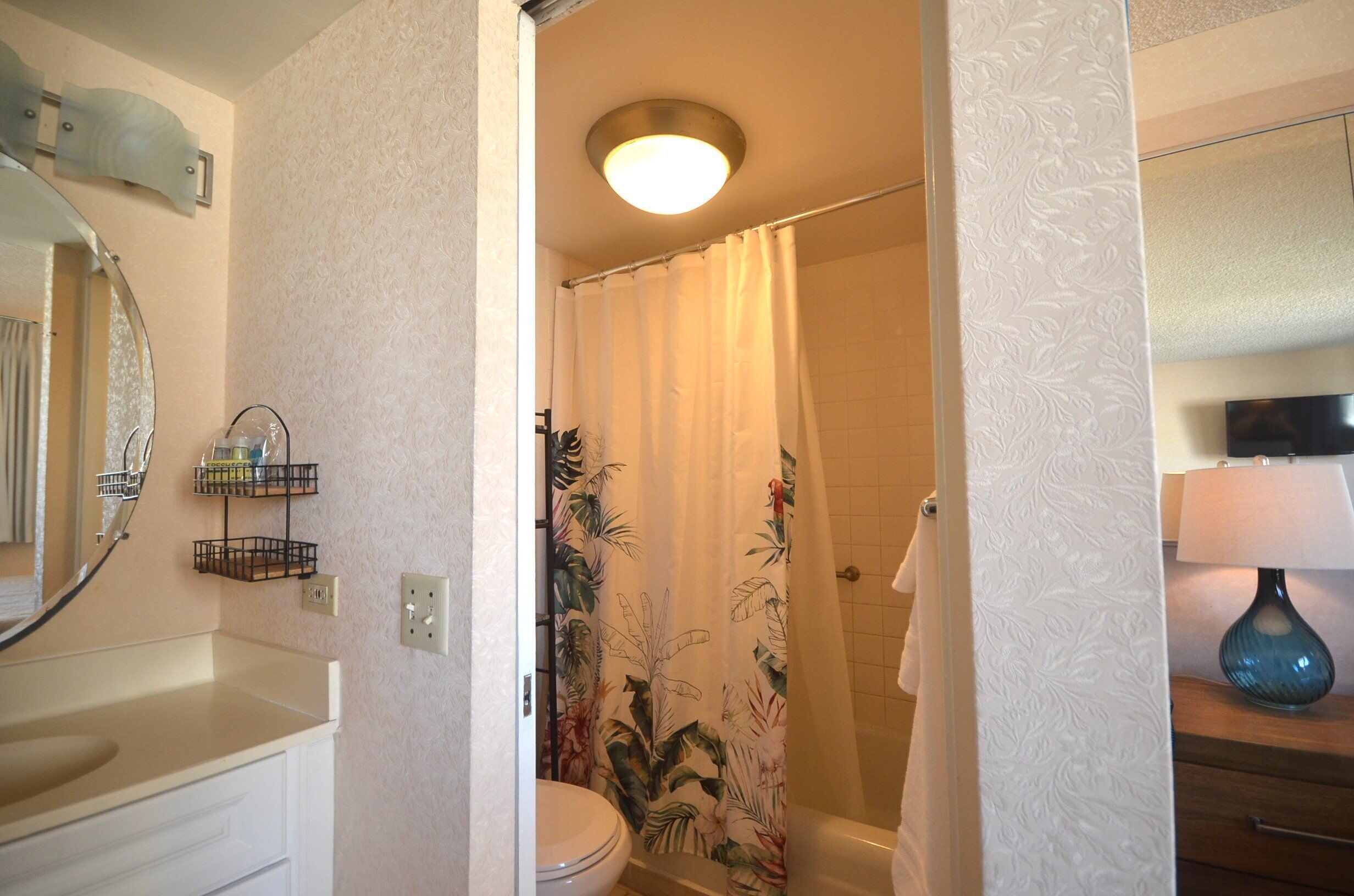 Une salle de bain comprenant des toilettes, un lavabo, un rideau de douche à motifs floraux, une serviette accrochée au mur et un miroir avec un luminaire placé au-dessus.