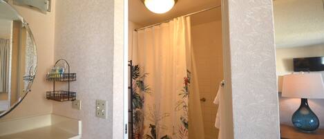 Une salle de bain comprenant des toilettes, un lavabo, un rideau de douche à motifs floraux, une serviette accrochée au mur et un miroir avec un luminaire placé au-dessus.
