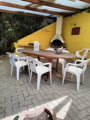 Een terras buiten met een houten tafel en witte plastic stoelen, een bakoven en een gele muur met een bord.