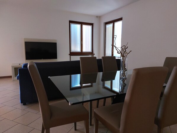 A modern living room with a glass dining table, beige chairs, a blue sofa, a flat-screen TV, and large windows with curtains.