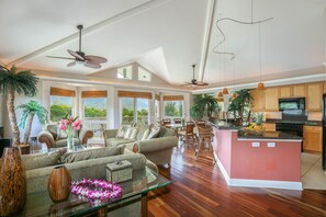 A spacious living area with a large sofa, a glass coffee table, and a dining area with chairs. There are ceiling fans, large windows, and a kitchen with wooden cabinets. - Nohea Villa: Elegant Tropical Home w/ Mountain Views, Pool & Spa (Princeville)